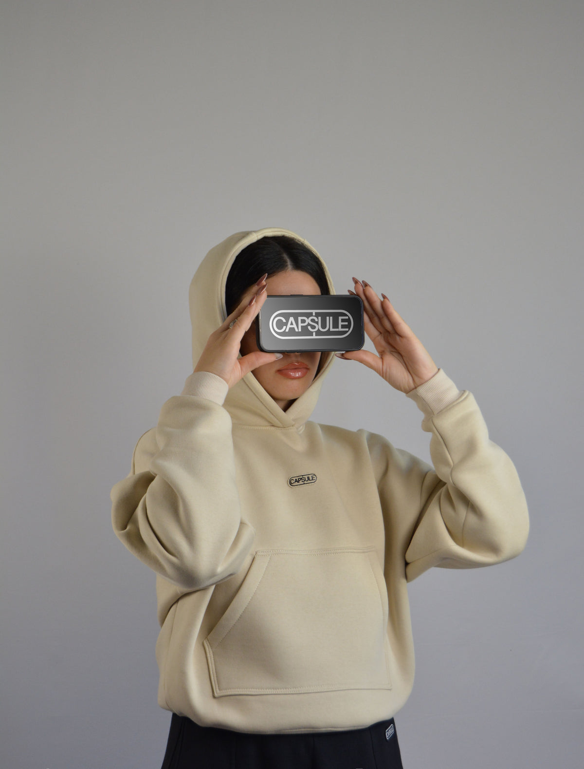 Model wearing a basic hoodie with a phone displaying 'CAPSULE' brand.